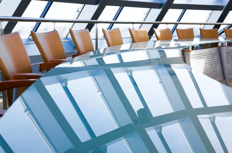 Modern conference room with curved glass table reflecting large windows and tan leather chairs.