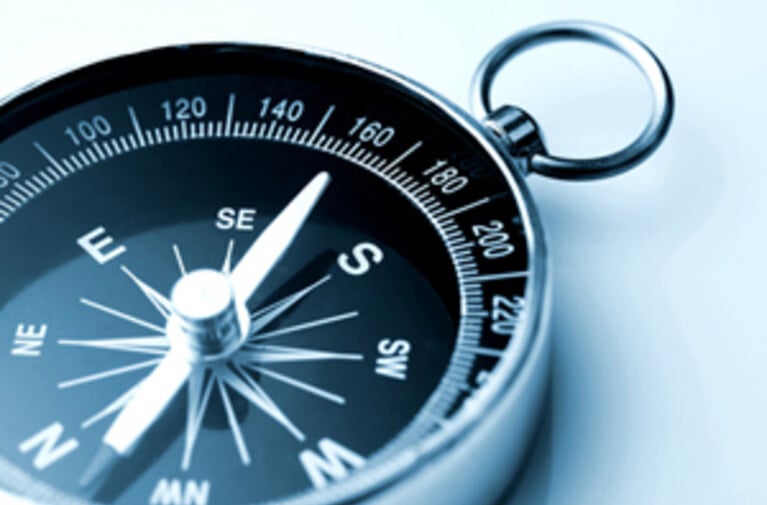 Close-up of a metallic compass showing cardinal directions and degree markings, with a silver ring, on a light background.